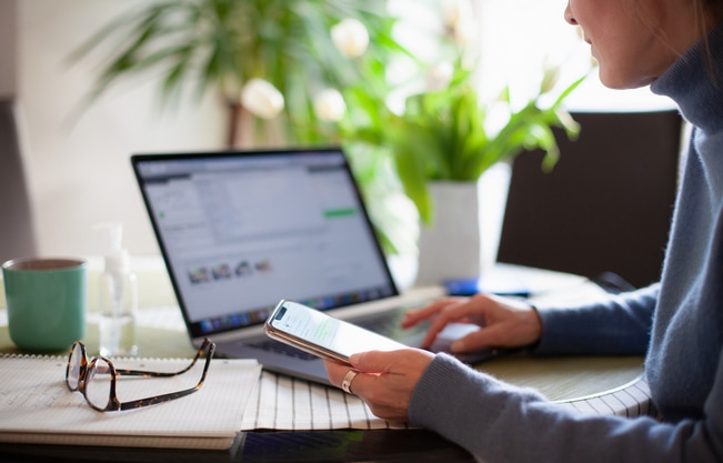 Woman working from home using laptop computer while reading text message on mobile phone