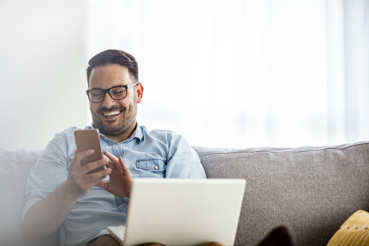 Smiling casual freelancer using smart phone and laptop while sitting on the sofa.