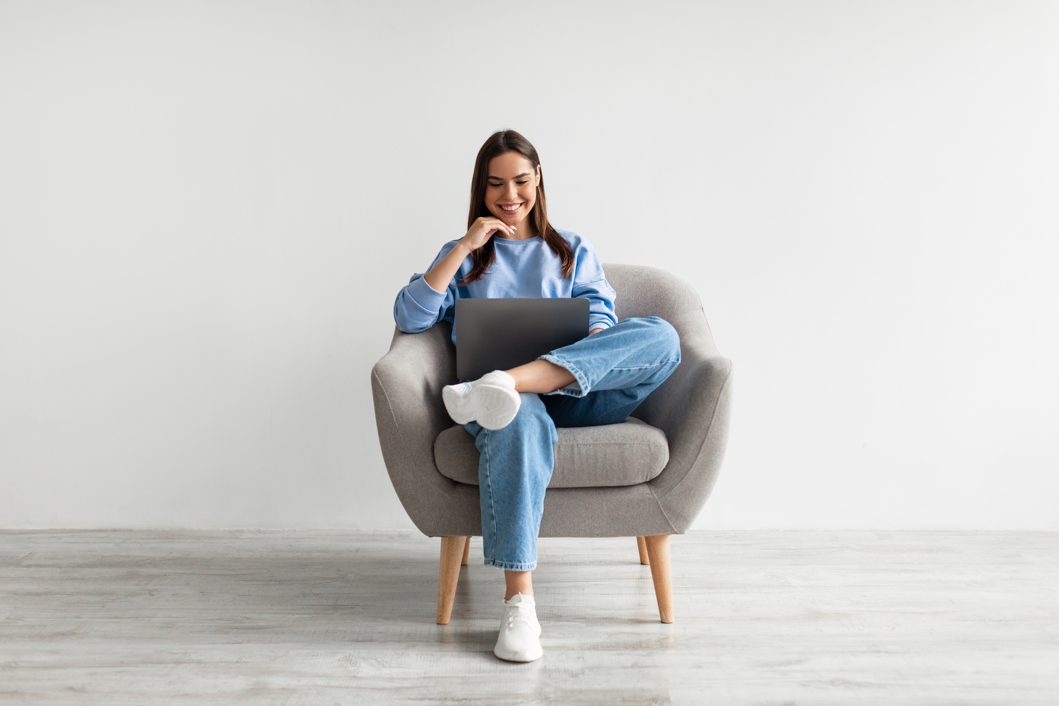 Millennial Caucasian lady in casual wear using laptop, sitting in armchair, having online conference against white wall