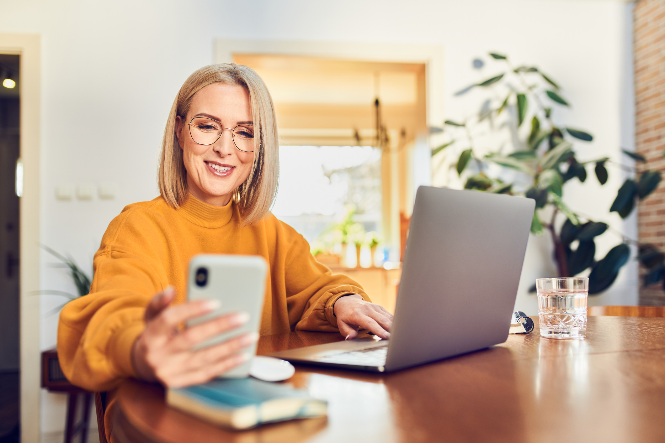 Smiling mature woman using smartphone working at home office with laptop sitting at dinning table